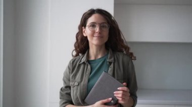 Kıvırcık saçlı, gözlüklü, esmer kadın ofis çalışanı elinde kağıt defterle kameraya doğru gülümsüyor.