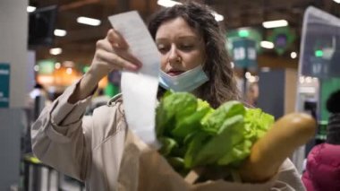 Tıp maskeli kadın süpermarketten alışveriş yaptıktan sonra çeke bakarken şok oluyor. Bayan tüketici alışveriş merkezinde elinde poşetlerle bekliyor.