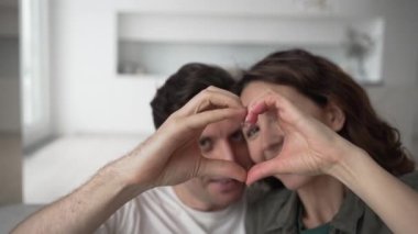 Happy couple in love showing heart shape with fingers and looking through it while sitting on sofa at home in living room