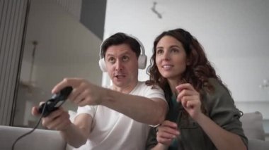 Excited young woman supports and kisses on cheek husband in headphones playing online games with remote controller in living room