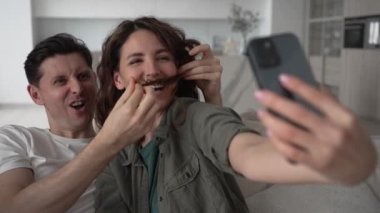 Laughing husband puts young wife long curly locks as theatrical mustache and poses against kitchen for funny videos and selfies closeup