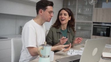 Happy married couple of young dark-haired man and curly woman discusses apartment renovation issues on paper drawings and laptop in kitchen