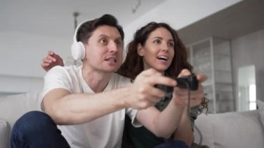 A man plays a video game with headphones and a gamepad in his hands on the couch at home, his wife sits next to her and supports her husband, cheers and worries about him and rejoices in the victory.