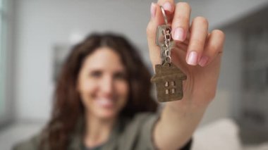 Happy smiling woman with brunette hair demonstrates key to new apartment with house key-chain against blurry kitchen extreme closeup