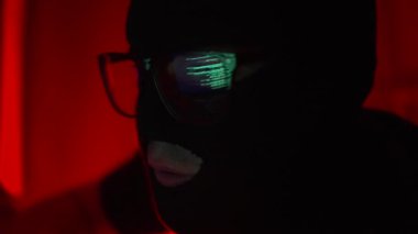 Hacker in a black mask and glasses sits in a dark room with a laptop and hacks into websites and databases