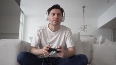 Excited man in wireless headphones sits on sofa in living room plays non-stop catch-up game with controller and loses illuminated by screen light