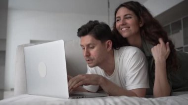 Joyful young couple of wife and brunet husband discusses chosen online purchases at sell-out via laptop lying on soft sofa in living room