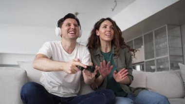Newlyweds of worried brunette woman and excited man in wireless headphones sit on sofa happy from win in play station game
