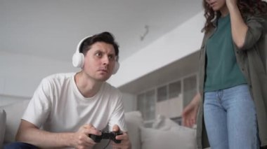 Worried wife comes up to husband in white wireless headphones playing computer games enthusiastically and quarrels and fights to stop player