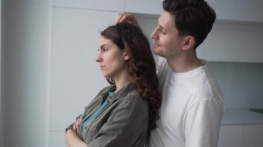 Young loving husband flirts with angry curly-haired wife standing by window after severe argument and woman forgives smiling closeup