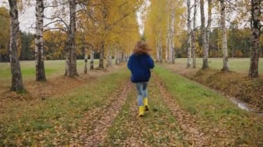 Rear view camera follows a woman in a blue jacket and yellow boots runs along a birch autumn alley in slow motion, the girl runs through the autumn park between yellow birches along the fallen leaves.