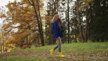 Woman in a blue jacket and yellow boots walks in the autumn park and enjoys the beauty, side view. Woman with curly hair collected a bouquet of yellow maple leaves.