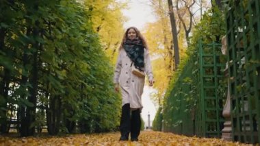 Pensive woman in a gray raincoat and wrapped in a scarf walks through the autumn park strewn with yellow leaves in slow motion