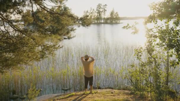 一个头戴草帽 身穿宽松衬衫的男人站在森林中荒岛的海岸上 夕阳西下时遥望远方 — 图库视频影像