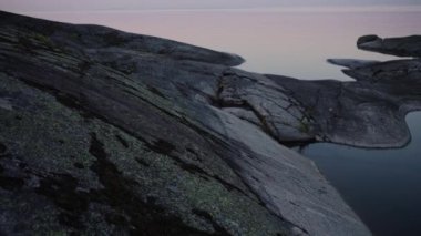 Gün batımında sakin bir göl üzerindeki taş sahilin manzarası. Soluk pembe gökyüzüne karşı pürüzsüz bir taş kıyısı.