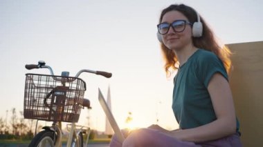 Genç bir kadın serbest çalışmaktan ve kendi zaman çizelgesine sahip olmaktan hoşlanıyor. Beyaz kulaklık takan iş kadını gün batımında bisiklet parkında oturuyor.