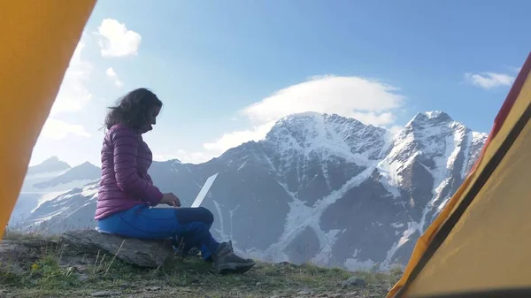 自由撰稿人的女人在高山间使用笔记本电脑 有着美丽的风景 一位观光客妇女坐在山中营地的石头上工作 从帐篷里看到概念 — 图库照片
