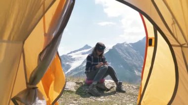 Sportif bayan yürüyüşçü, yaz mevsiminde yüksek dağlarda bir çadırın içinde, bir kayanın üzerinde kamp yaparken akıllı telefon kullanır.