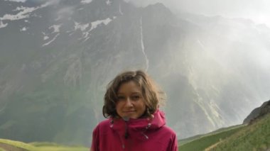Dağlarda sağanak yağış altında duran bir kadın yürüyüşçünün portresi, yaz mevsiminde yeşil dağların arka planına karşı pembe zarı ceketli bir yürüyüşçü, soğuktan ve gülümsemelerden çekiniyor, yavaş çekim.