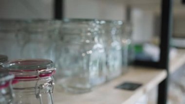 Kamera, bir eco dükkanında ambalajsız cam şişelerle rafta hareket ediyor, bir kadının eli raftan bir kavanoz alıyor, kamera hareketi. Bilinçli tüketim ve endişe