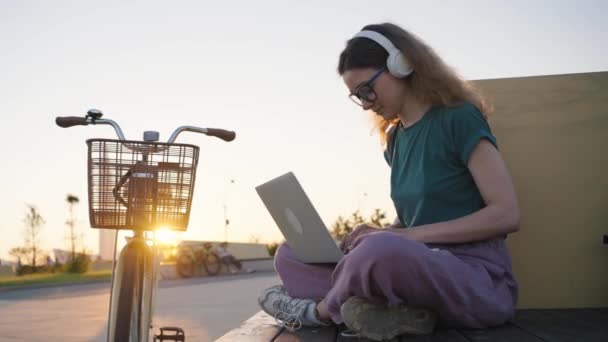 在城市公园的长椅上 一个戴眼镜的女人在笔记本电脑上远程工作 一个拿着自行车的学生在户外学习 享受日落的美景 慢动作 — 图库视频影像