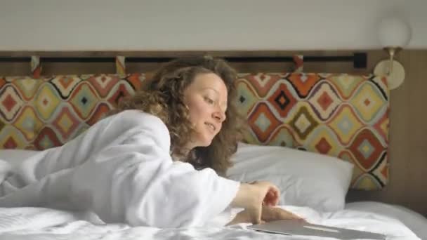 Щаслива Кучерява Жінка Білому Халаті Розслабляється Ліжку Ноутбуком Турист Готелі — стокове відео