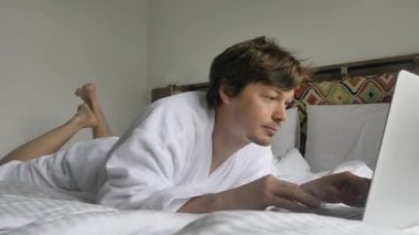 Beyaz bornozlu, mutlu, yakışıklı bir adam göbeğiyle yatağa uzanır ve yüzünde bir gülümsemeyle güler ve bilgisayardaki bir mesaja cevap verir, klavyeye mesaj yazar.
