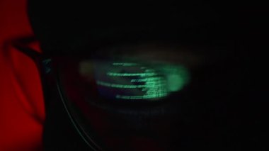 Siyah maskeli ve gözlüklü bir hacker bir sunucu veya siteyi hacklemeye çalışıyor. Kırmızı arka plandaki gözlüklerin lenslerindeki kodun yansıması.