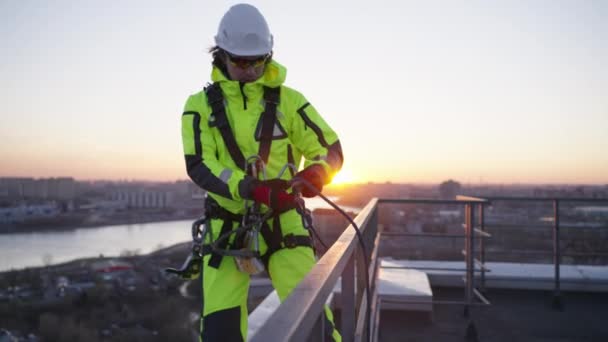 産業用クライマーは屋根の端に立っており 日没の背景を背景に建物から降りるためにロープを結びます 標高の高い仕事 危険な極端な仕事 — ストック動画