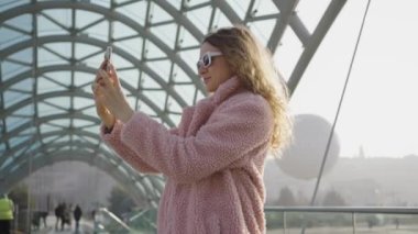 Pembe kürklü ve gözlüklü genç bir kız güneşli bir günde modern cam bir köprüde akıllı bir telefondan selfie çekiyor. Kadın turist Tiflis 'te yürüyor