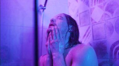 Uzun saçlı bir adam banyodaki sıcak su deresinin altından kalkar ve kendini neon pembe ve mavi ışıkla kaplamaya başlar.