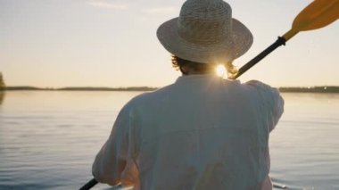 Hasır şapkalı bir adam gölün sakin yüzeyindeki kanoda gün batımına doğru kürek çeker. Yürüyüşçü sakin açık hava aktivitelerinin tadını çıkarıyor..