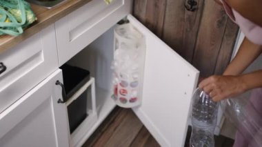 Evde ayrı ayrı çöp toplayan bir kadın, mutfaktaki lavabonun altındaki bir kutunun içindeki plastik şişeleri atık geri dönüşüm kutularına koyuyor. Çevreye önem vermek, uzayın ehil dağılımı