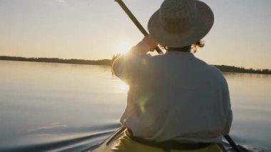 Hasır şapkalı ve keten gömlekli bir adam, suyun pürüzsüz yüzeyinde bir spor kanosunun içinde yüzüyor, yazın günbatımında sakin bir şekilde kürek çekiyor, canlı kamera.