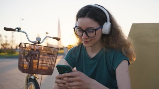 在夏天的特写中 穿着眼镜和耳机的可爱微笑的女人坐在城市广场上 骑着自行车 一边用手机 千禧年女人用智能手机交流 — 图库视频影像