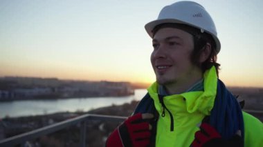 Gün batımında teçhizatlı gülümseyen endüstriyel bir tırmanışçının portresi ve beyaz bir şapkayla omuzlarında bir iple çatıda duran bir işçi.