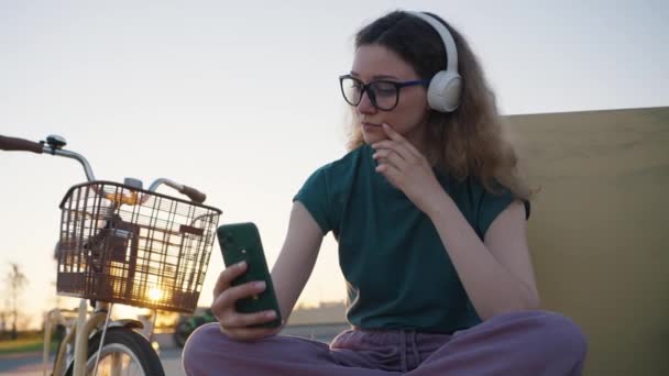 千年紀の少女は携帯電話を手に公園のベンチに座って セフィを作り 日没時にワイヤレスヘッドフォンで音楽を聴く 暖かい夏の夜に市内公園をサイクリング — ストック動画