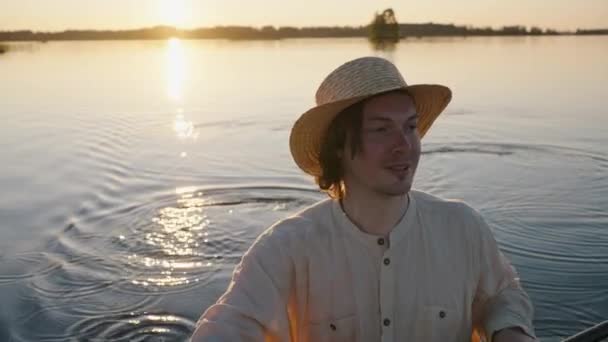 夕暮れ時の夏にはカヤックを漕ぐ藁帽子とリネンのシャツを着た映画人 湖の後ろに沈む太陽 コンセプト — ストック動画