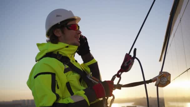 在落日的背景下 一名高空工作者正在用悬挂在大楼正面 系在安全带上的手机与降落者交谈 一个工业登山者在 — 图库视频影像