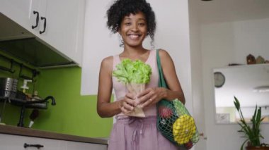 Mutlu Afrikalı Amerikalı kadın mutfakta elinde sebze, elinde salata ve gülücüklerle tekrar kullanılabilir bir çantayla duruyor. Bilinçli tüketim kavramı, sıfır atık, çevreye özen