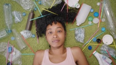 Plastik şişeler ve geri dönüşüm, karışık ırk kıvırcık kadın plastiğin arasında yeşil bir halıya uzanır ve gözünün üstüne içki şişelerinden kapaklar koyar, hayali bir solaryumda bronzlaşır veya güneş altında gözlüklü ve