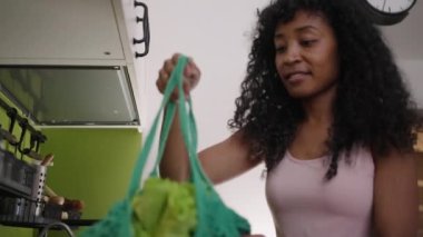Afro-Amerikalı bir kadın mutfaktaki yeşil bir ağ torbasından taze sebzeler çıkarıyor. Çevre dostu bir çiftlik dükkanından taze otlar alıyorum..