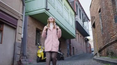 Pembe kürklü ve güneş gözlüklü şık bir bayan turist sırt çantasıyla etrafta dolaşıyor ve binaya bakıyor, mimarinin güzelliğine hayran, gezgin bir kadın