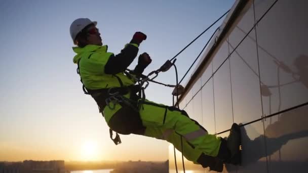 在日落时分 一位工作着的工业登山者在一幢多层楼房的立面上挂在绳索上 一边笑一边跳舞 那人挥挥手 竖起大拇指 — 图库视频影像
