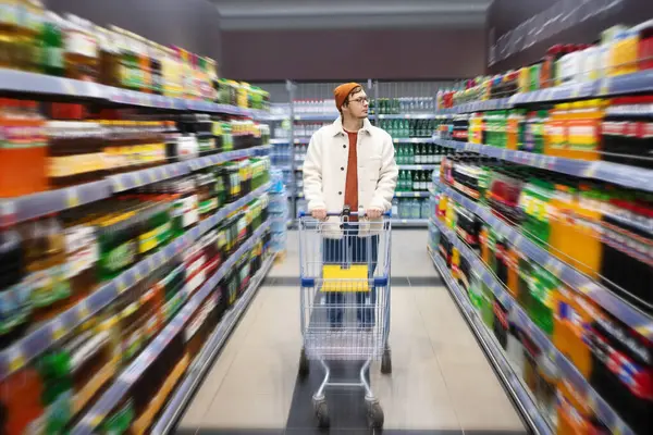 A man stands with a shopping cart in a supermarket between the shelves with drinks and chooses juice. A stylish man chooses products in a grocery store.