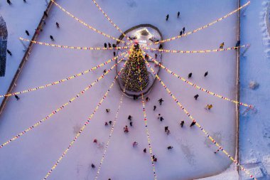 Akşamları paten sahasının ortasında çelenkler ve bayraklarla süslenmiş Noel ağacı, hava manzaralı. İnsanlar parkta buz pateni pistinde kayıyorlar, neşeli bir atmosferde..