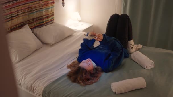 一个女人躺在床上 一边高高地拿着电话 一边和朋友聊天 一边在晚上上网购物 一个女孩在漫长的一天工作之后 正在轻松地翻阅新闻 — 图库视频影像