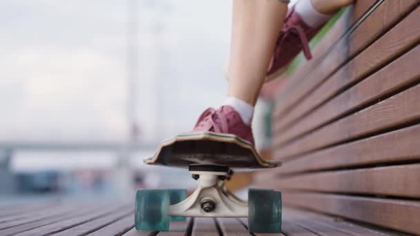 少女坐在街上的木制长椅上 脚踏在滑板上 年轻女孩过着积极的生活 做着极端的运动 — 图库视频影像