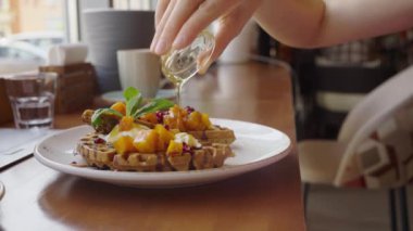 Bir insan eli, doğranmış mango, nar tohumu ve beyaz bir tabakta servis edilen nane yaprağı ile kaplı yeni hazırlanmış bir waffle 'ın üzerine şurup döküyor. Vitamin zengini sağlıklı ve lezzetli bir kahvaltı.