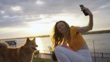 Pozitif kadın, kırmızı köpeğiyle birlikte gün batımının arka planına karşı sette yürürken akıllı telefonuyla selfie çekiyor. Bir kız dört bacaklı arkadaşıyla vakit geçirir..
